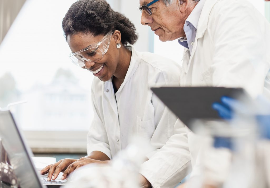 Two researchers in lab coats and goggles look together at a laptop scream. One is smiling.